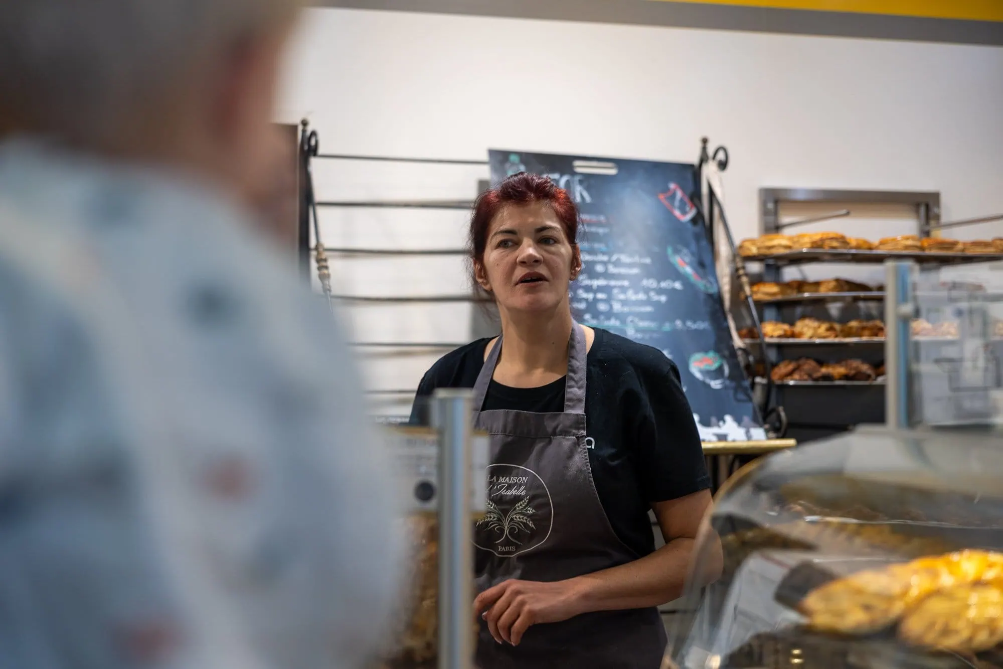 Frau mit roten Haaren in einer Schürze steht in einer Bäckerei zwischen Brotregalen