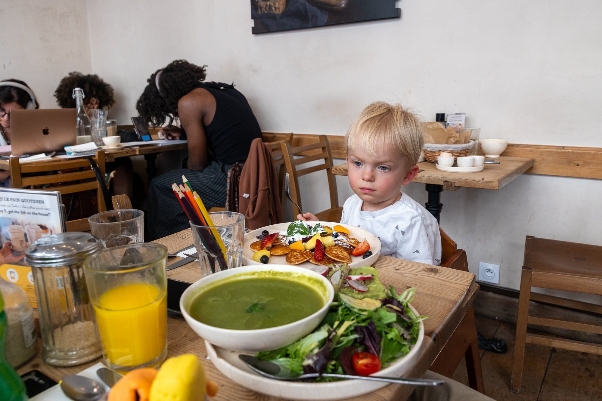 Ce să mănânci la Paris: Ghid foodie complet 2026 (bistrouri, Michelin, scena vegetariană) 17 Copil mic cu părul deschis stând la masă cu o farfurie de supă verde și clătite într-o cafenea