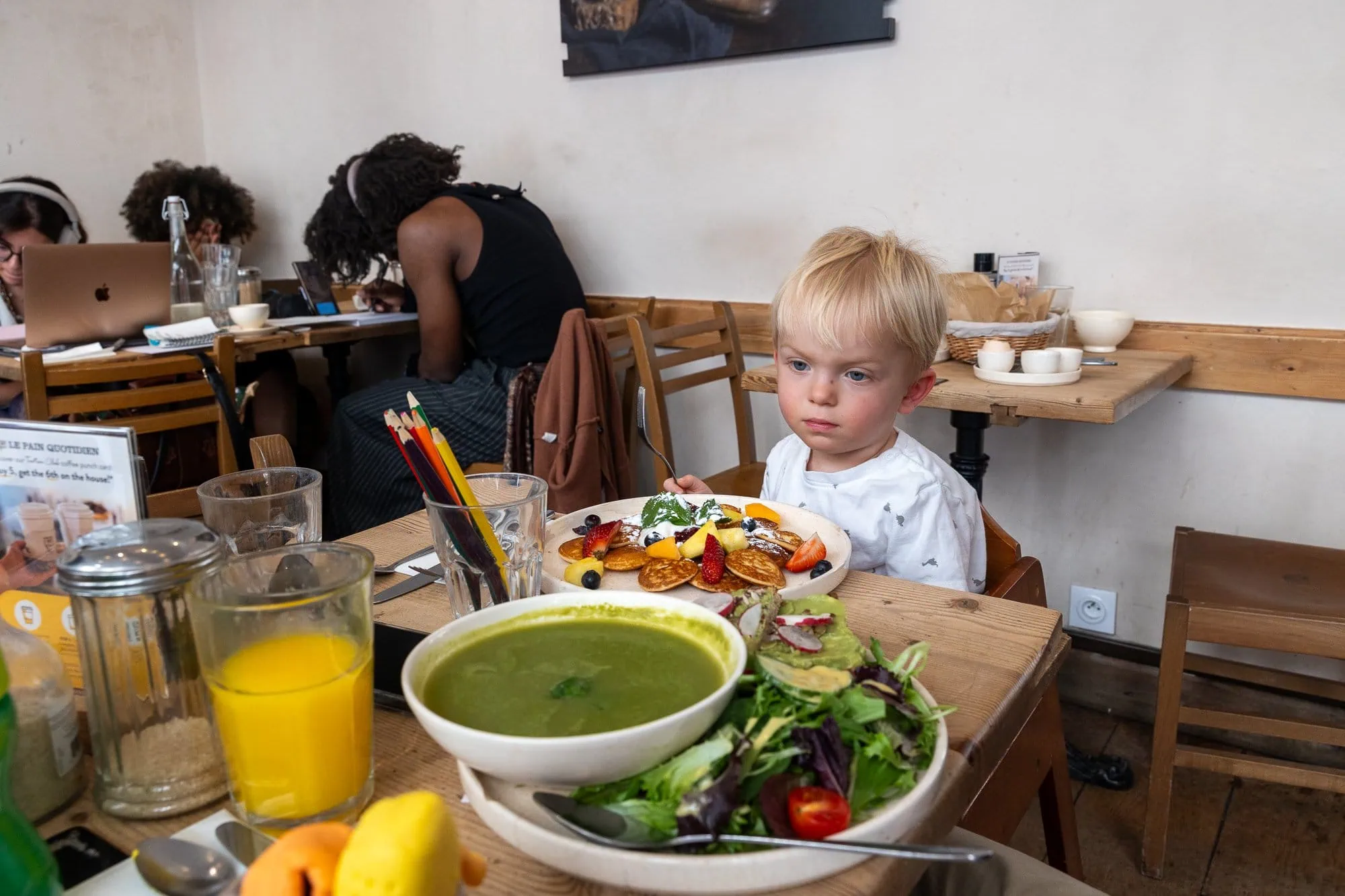 Kleines Kind mit hellen Haaren sitzt am Tisch mit einem Teller grüner Suppe und Pancakes in einem Café