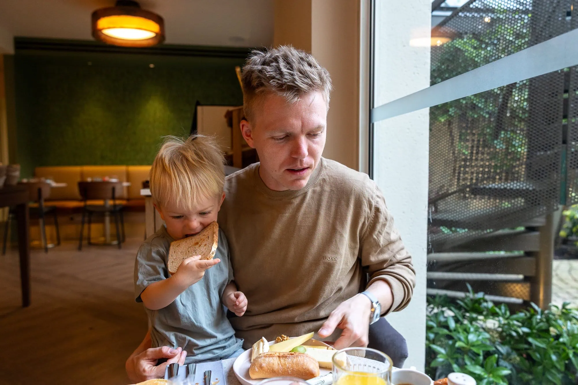 Mann in beigem Pullover hält ein kleines Kind, das ein Sandwich in einem modernen Café kaut