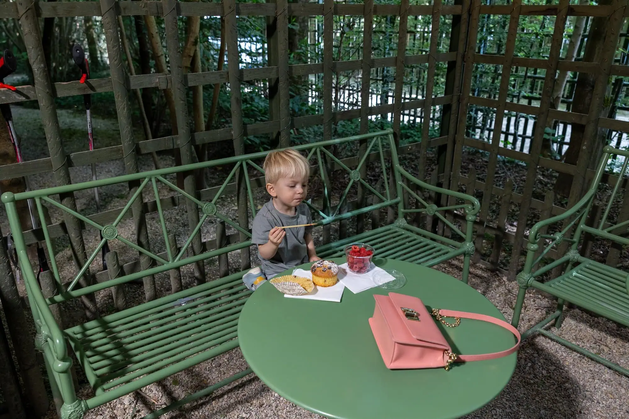Kleines Kind sitzt auf einer grünen Bank in einem Pariser Gartencafé mit Süßigkeiten