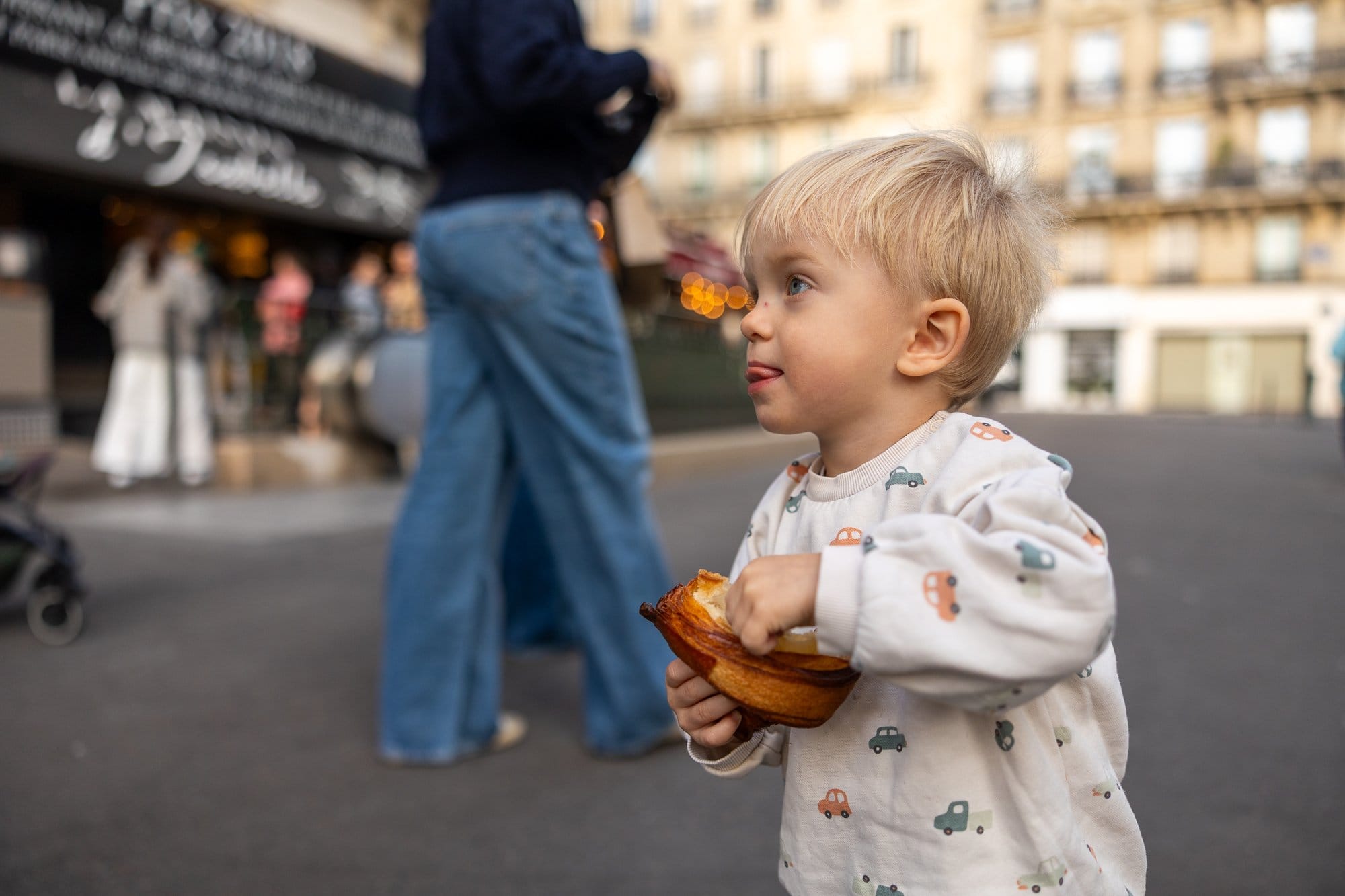 Ce să mănânci la Paris: Ghid foodie complet 2026 (bistrouri, Michelin, scena vegetariană) 11 Băiețel blond în tricou alb cu mașinuțe ținând o bucată de brânză pe o stradă pariziană