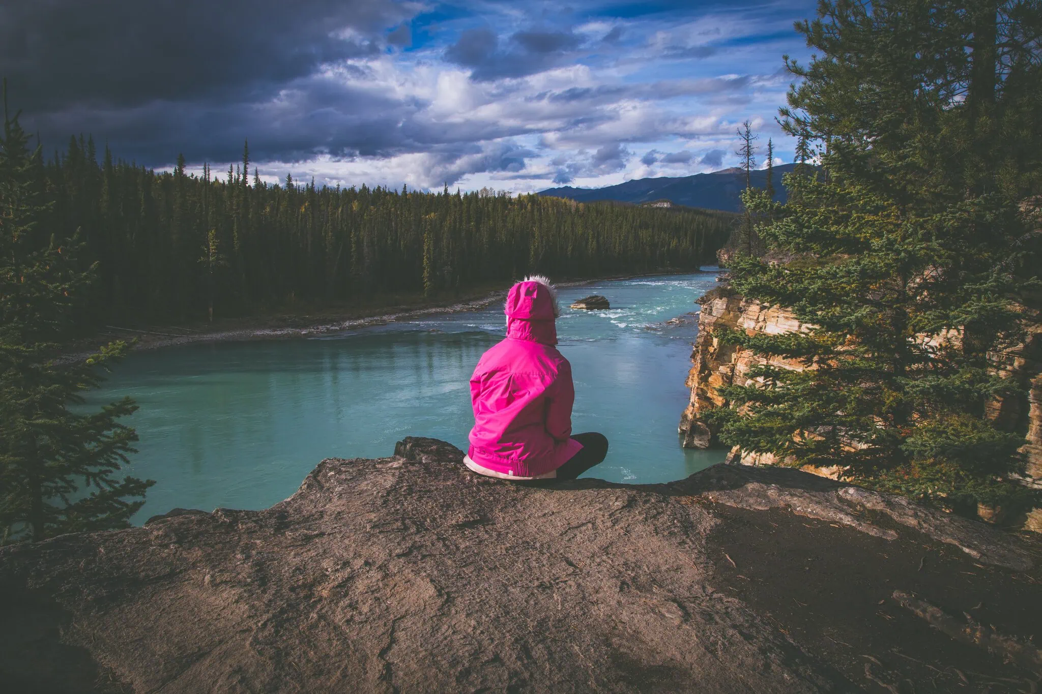 Lucie sitting in pink on a cliff above the turquoise canyon of Athabasca Falls