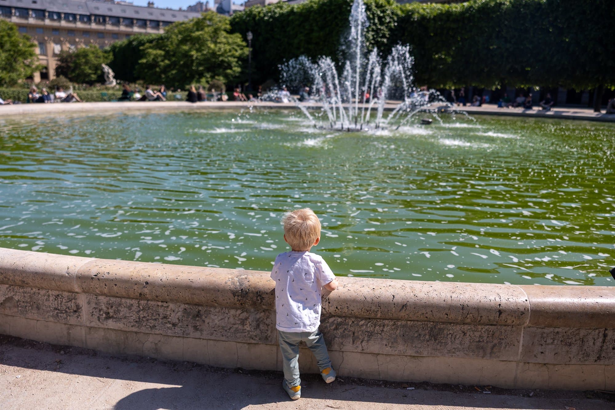 Paris cu copii: Ghidul complet pentru un city break în familie (la ce să te aștepți cu căruciorul) 5 Un copil mic în tricou alb stă lângă un balustrat de ciment și privește o fântână într-o grădină pariziană