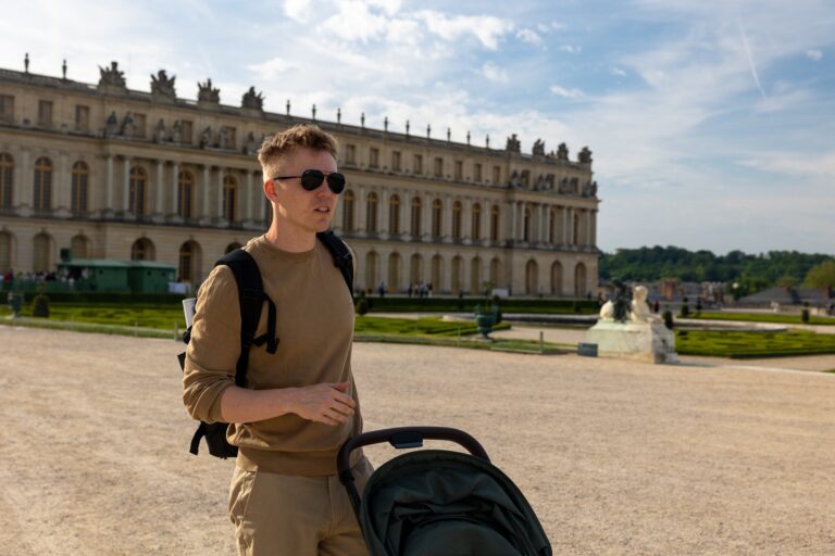 Muž v tričku s brýlemi stojí s kočárkem před Versailleským palácem