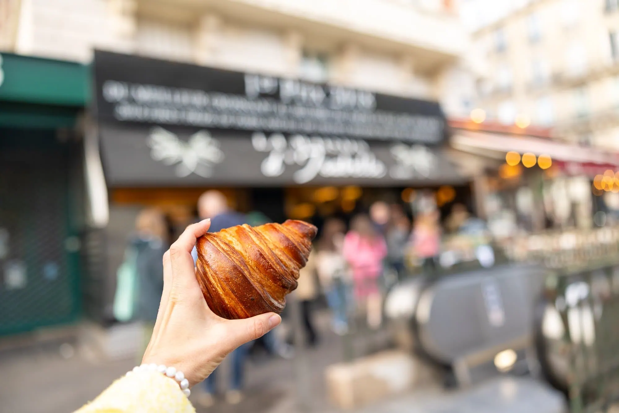Pařížská etiketa: Jak nebýt za burana (slovníček, pravidla, co NEdělat) 5 Ruka drží zlatavý croissant před pařížskou pekárnou s lidmi v pozadí