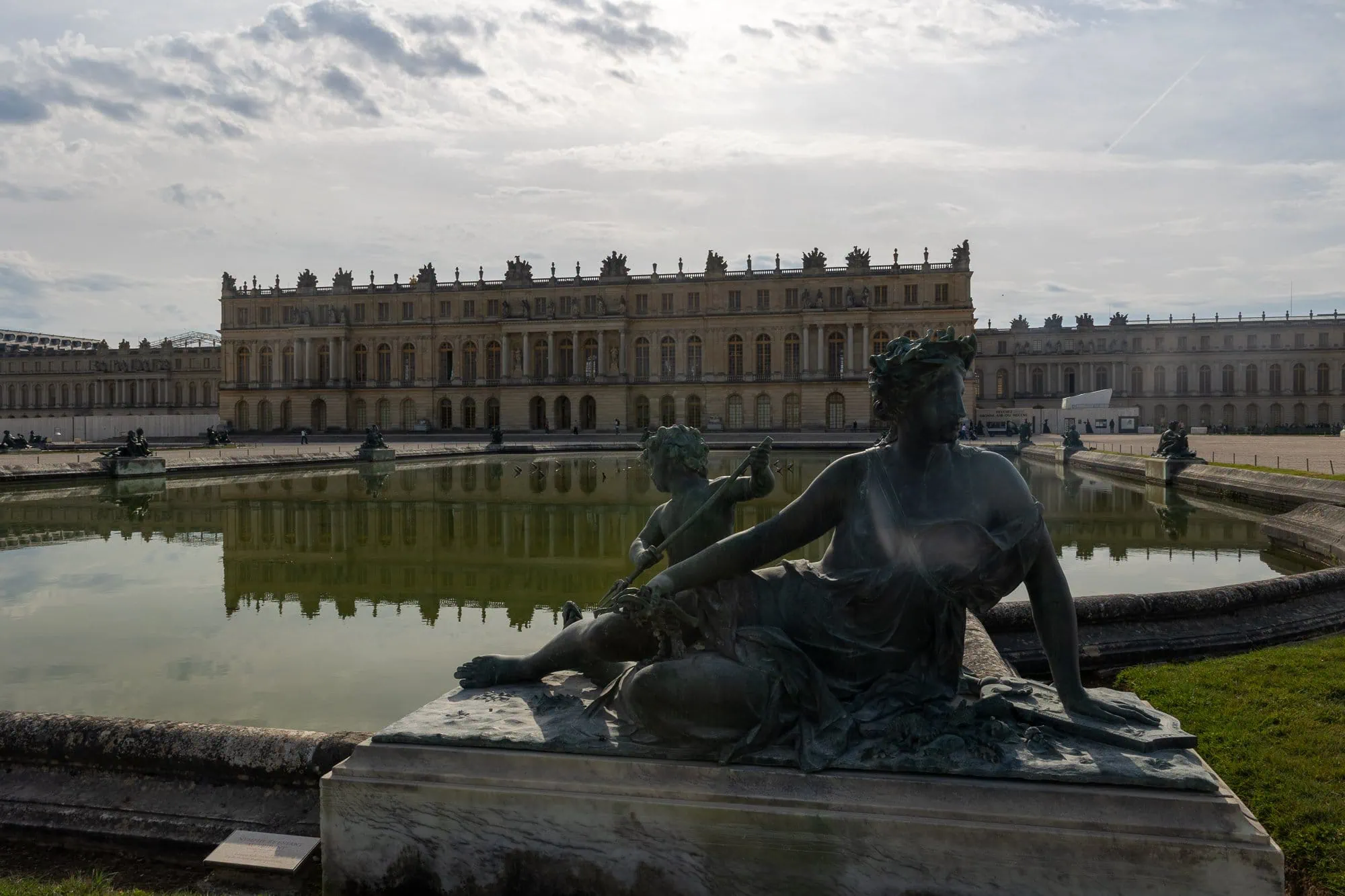 Bronzová socha muže v parku před Versailleským palácem s rybníkem